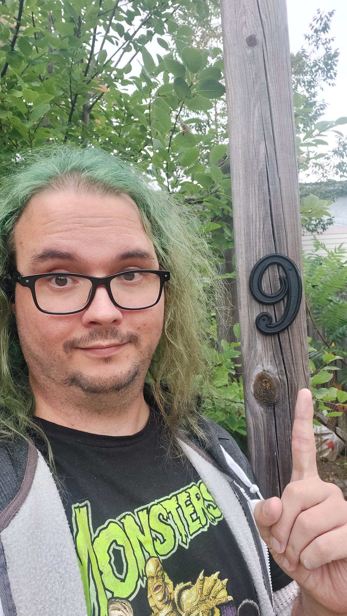 A man with long green hair and no beard stands in front of a post with a number 9 on it